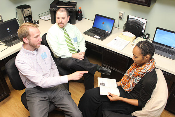 Three people sitting in an office engaged in a discussion. Two men, one pointing, and one woman holding an open book. Two laptops, papers, and office supplies are on the desk in the background.