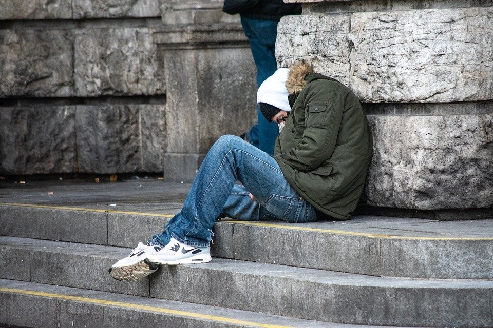 A person sits hunched over on stone steps, wearing a hooded green jacket, jeans, and white sneakers. A portion of another person's legs is visible behind them.