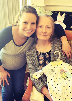 A smiling pregnant woman is standing next to an older woman who is seated and holding a baby outfit with a bee design.