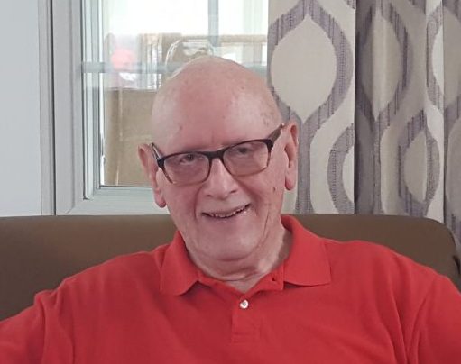 An elderly man with glasses and a red polo shirt sits in front of a window with patterned curtains, smiling at the camera.