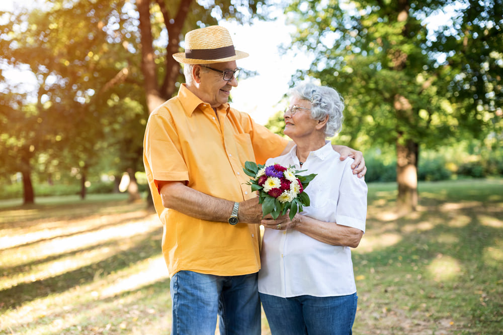 An elderly man and woman standing in a park, looking at each other and smiling. The man is wearing a hat and orange shirt, holding a bouquet of flowers. The woman wears a white shirt. Trees in the background.