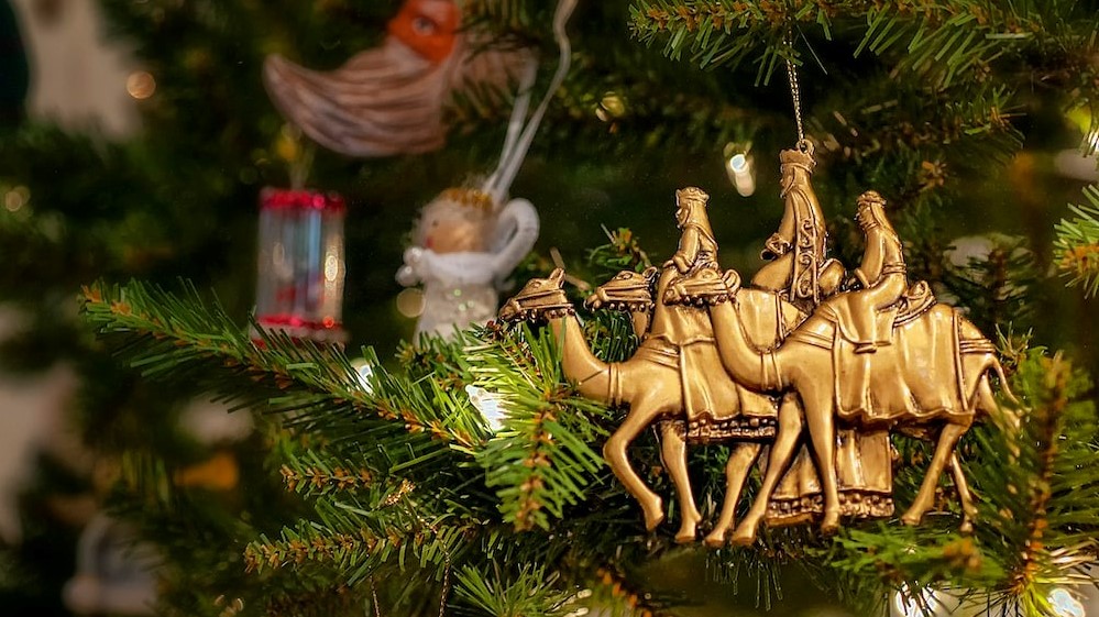 Close-up of a Christmas tree branch with an ornament depicting the Three Wise Men on camels, along with other festive decorations.