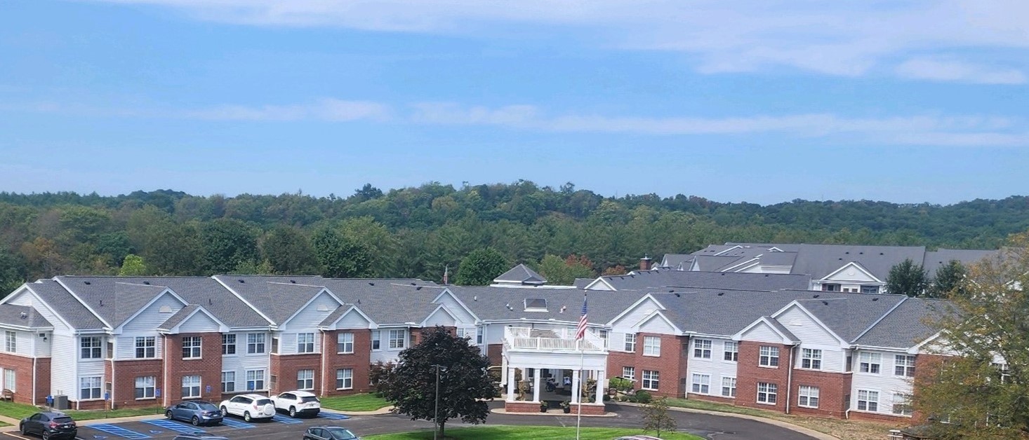 A large, two-story brick and white building with a prominent central entrance under a blue sky, surrounded by trees and greenery, hosts The Glenwood Community Entrepreneur in Residence program. Several vehicles are parked in front.