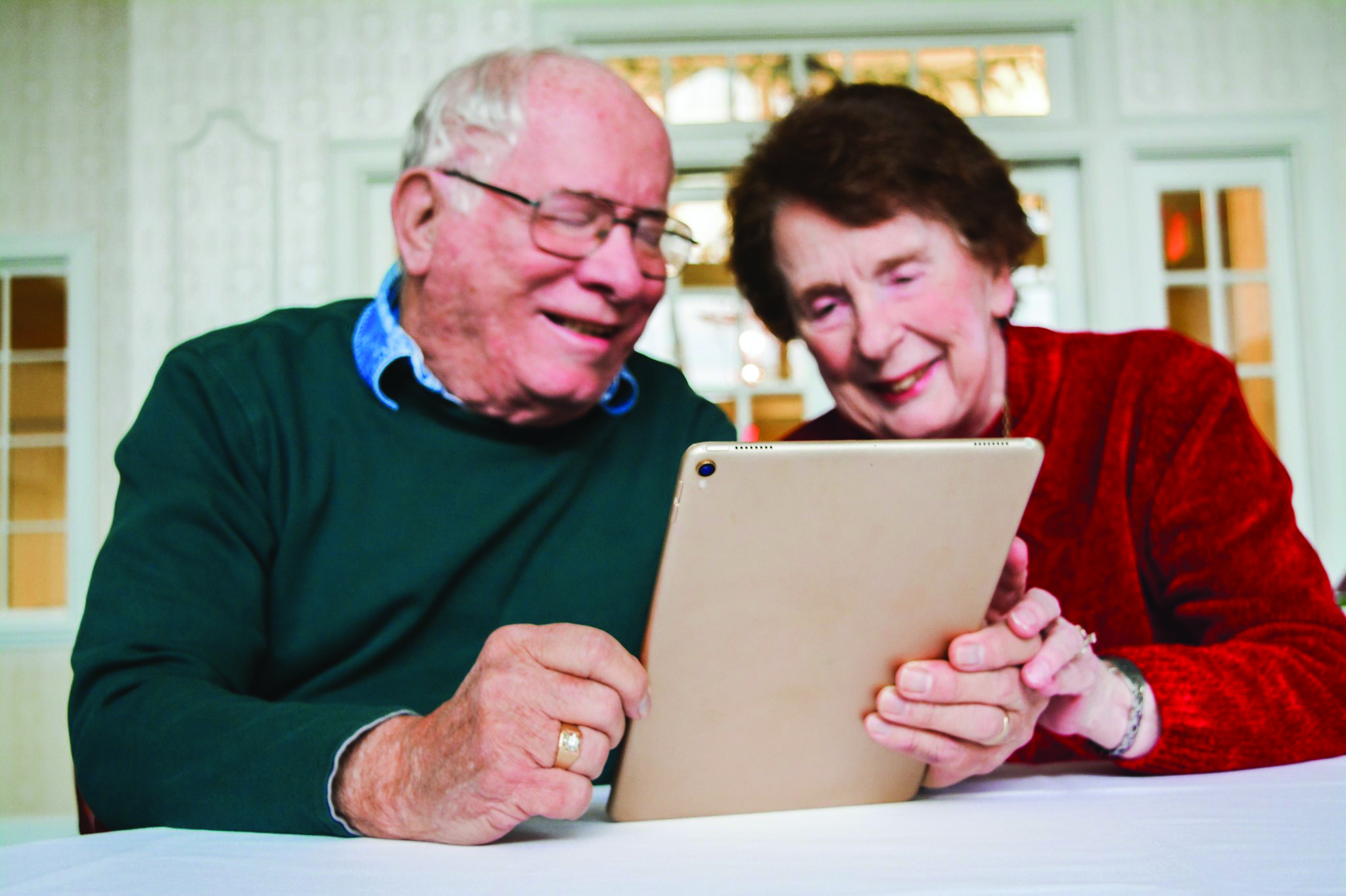 An elderly couple is engaging with technology, using a tablet together at a table in a bright room, smiling at the screen.