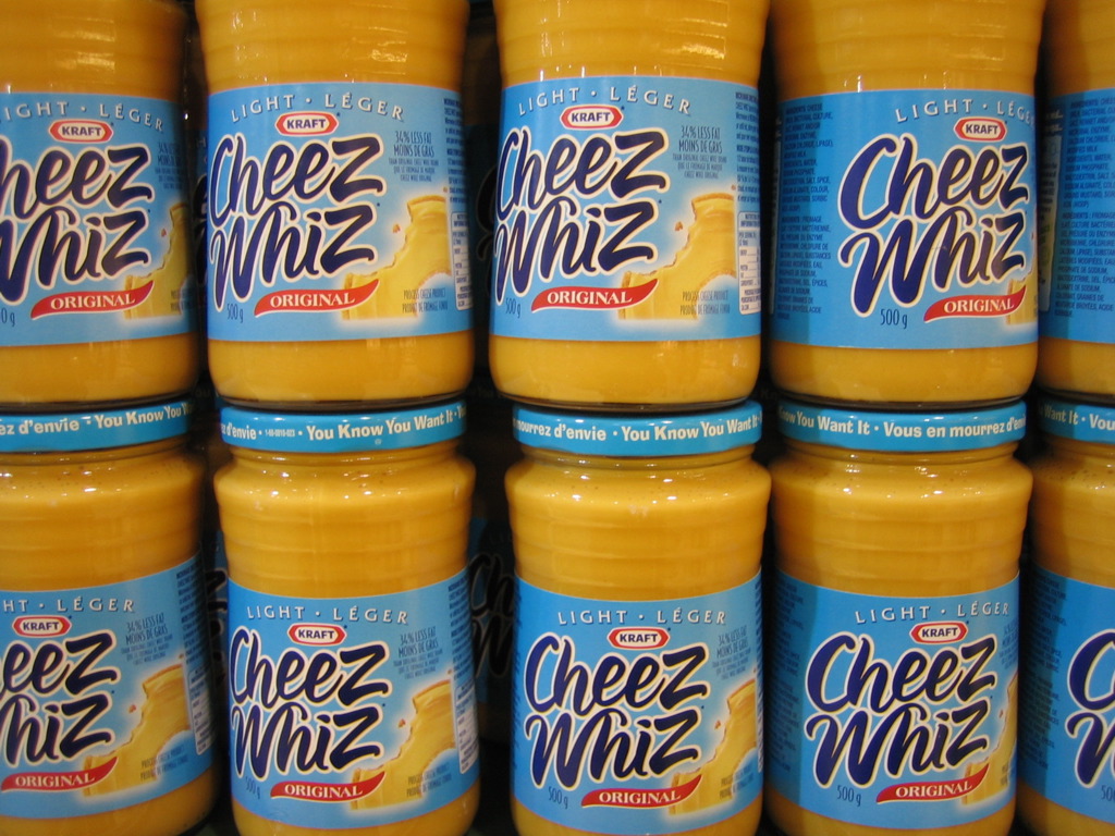 Stacks of jars of Kraft Cheez Whiz Light Original on a shelf, with blue labels and yellow product visible inside.