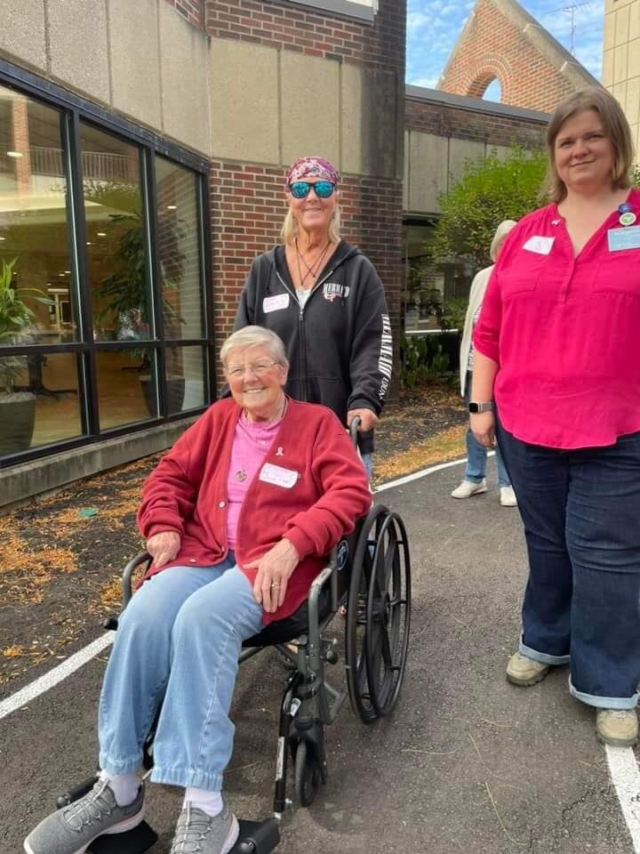 Three people outdoors; one elderly individual in a wheelchair, one standing behind, pushing the wheelchair, and another individual standing to the right. Both adults are wearing name tags representing their careers at United Church Homes.