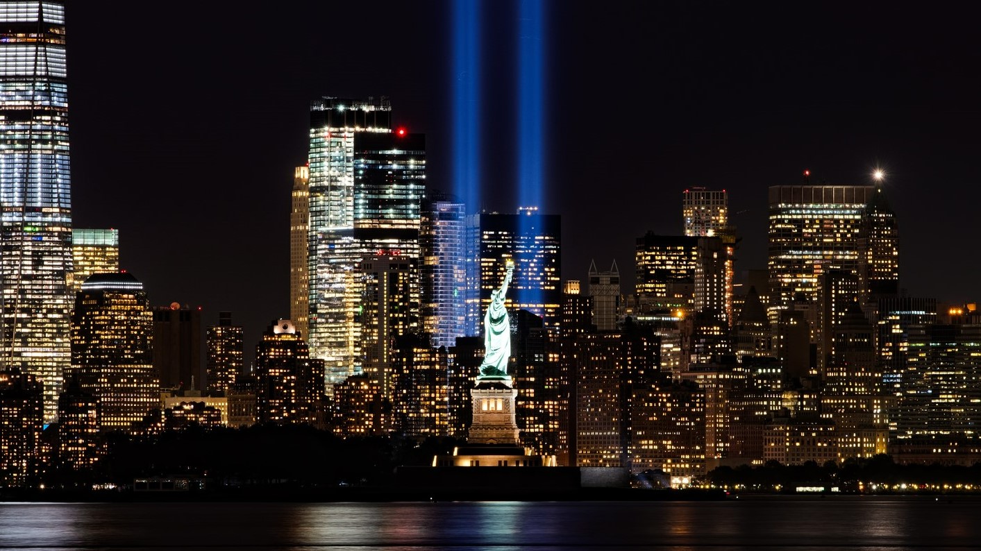 The Statue of Liberty illuminated at night with two blue beams of light extending into the sky, set against the New York City skyline.