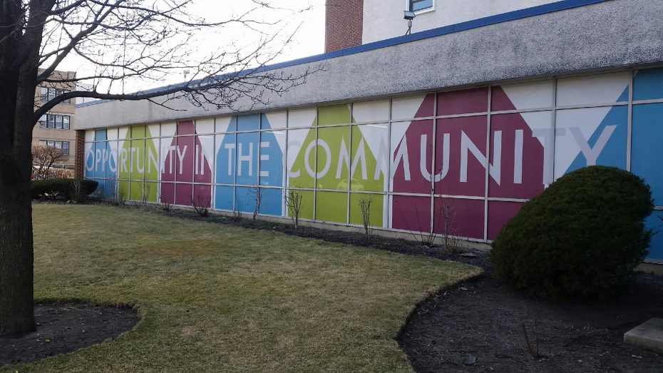 A building wall features colorful geometric shapes and the message "Opportunity in the Community." The foreground includes a lawn, shrubs, and a leafless tree.