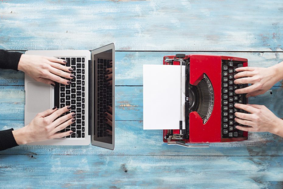 Two people typing, one on a laptop and the other on a red typewriter, placed on a blue wooden surface.