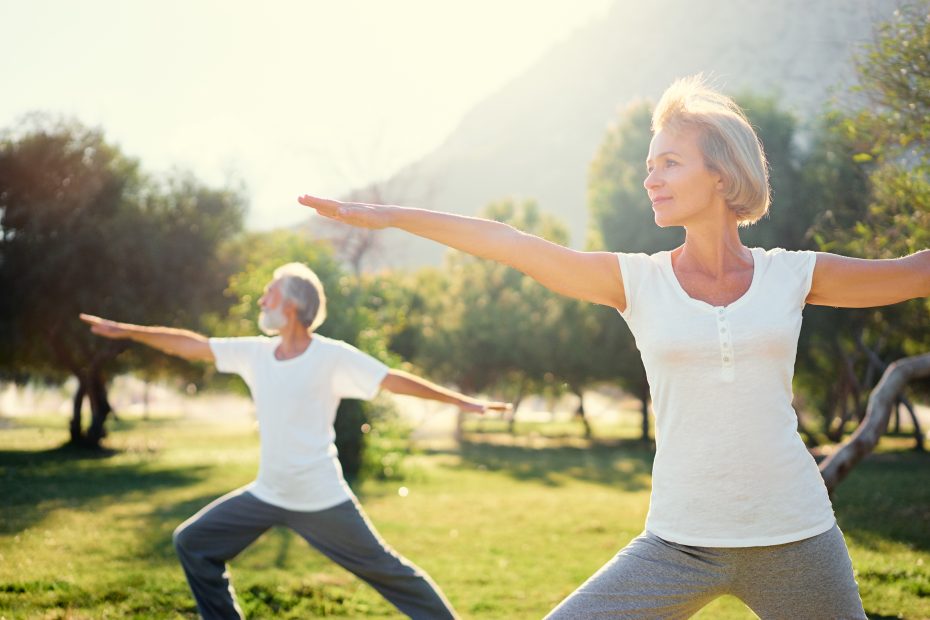 Two people, an older man and woman, are practicing yoga outdoors in a park. They are doing the Warrior II pose, standing with arms extended. The background includes trees and a sunlight-filled sky.