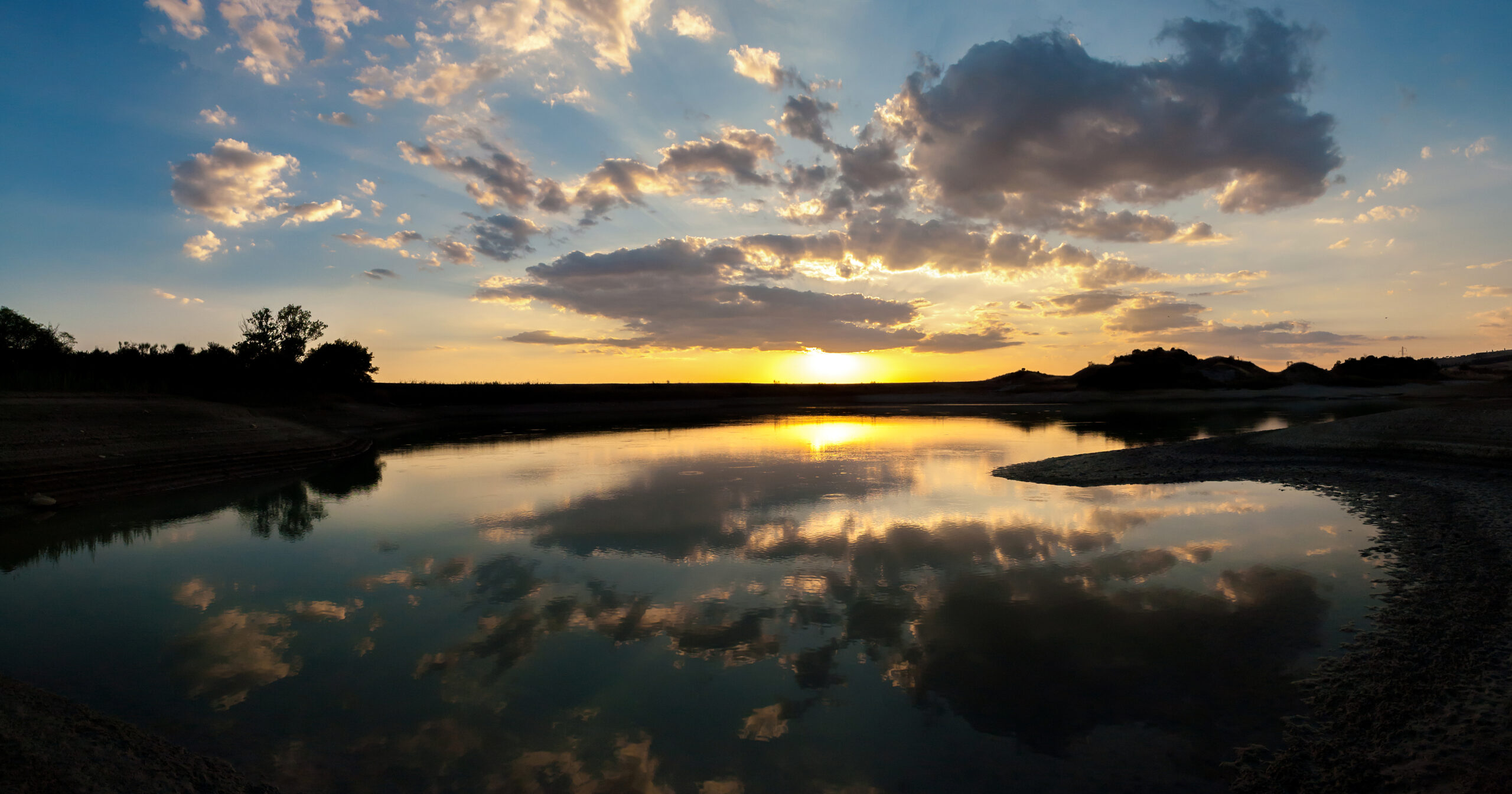 A serene lake at sunset with clouds reflected on the water's surface and trees silhouetted against the horizon.