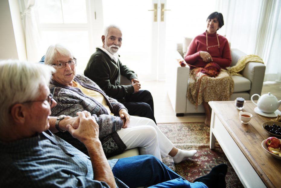 Four elderly people are seated in a living room engaged in conversation. Two men and a woman are on a couch, and another woman sits in an armchair. A table with snacks and drinks is in front of them.