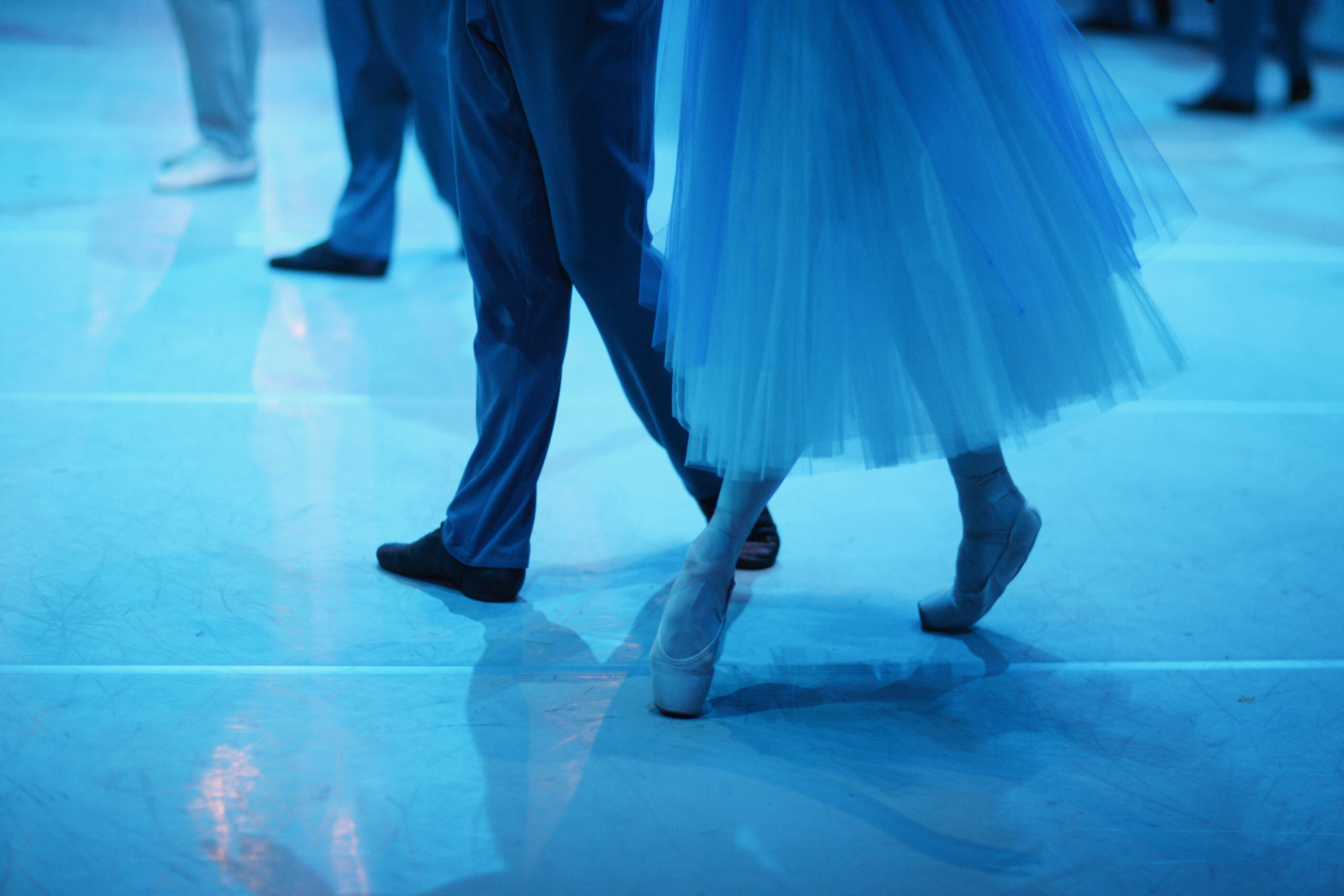 Two ballet dancers performing on stage, with the focus on their legs and blue costumes under blue lighting.