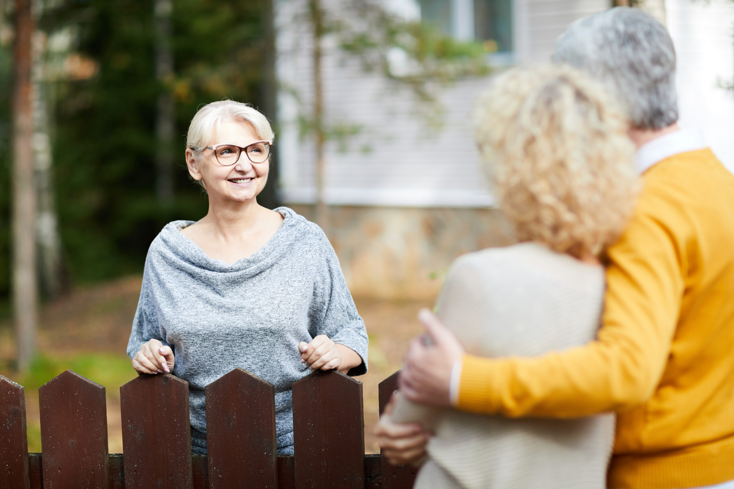 Older woman with glasses leans on a wooden fence, smiling and engaging with a couple who stand close together, facing her with their backs to the camera. Trees and a house are in the background.