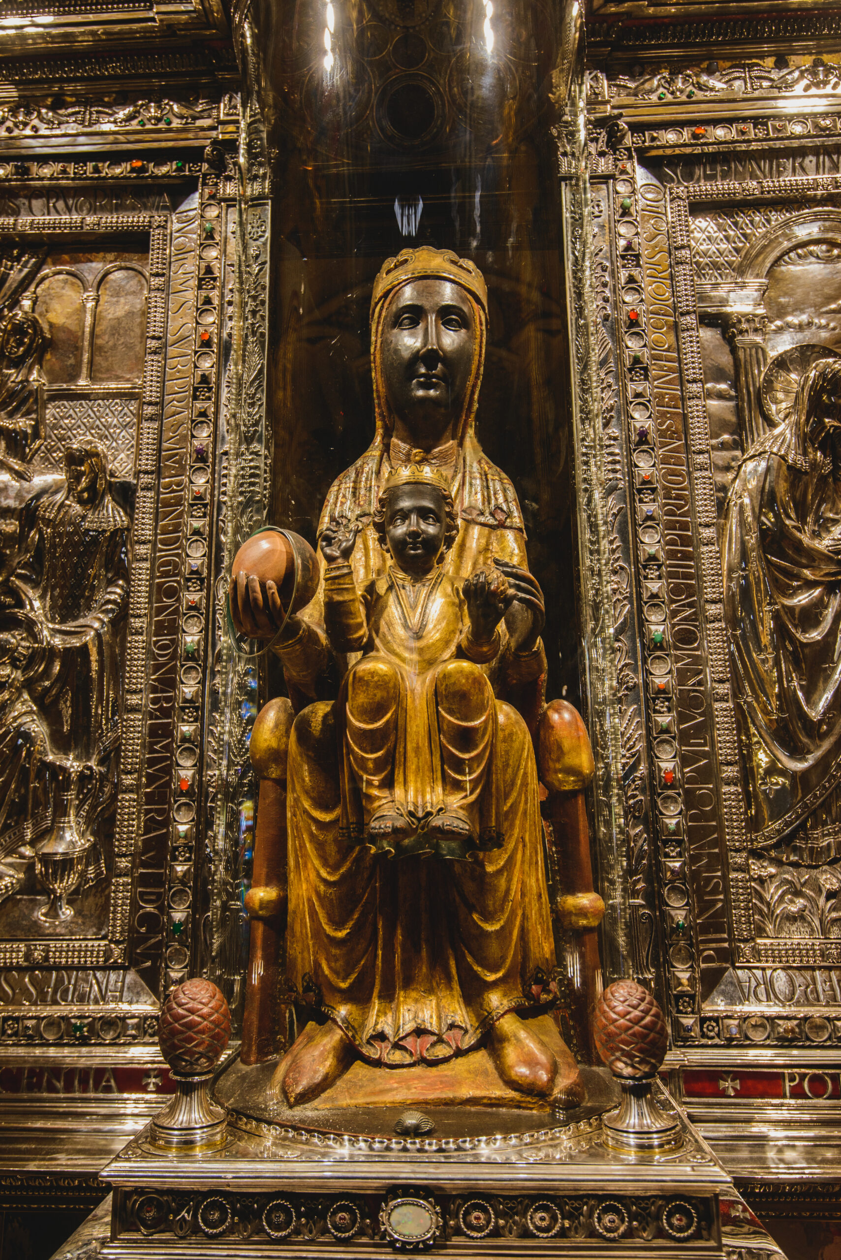 A golden statue of a seated woman holding a child, adorned with intricate details and set in an ornate background featuring religious carvings and gemstones.