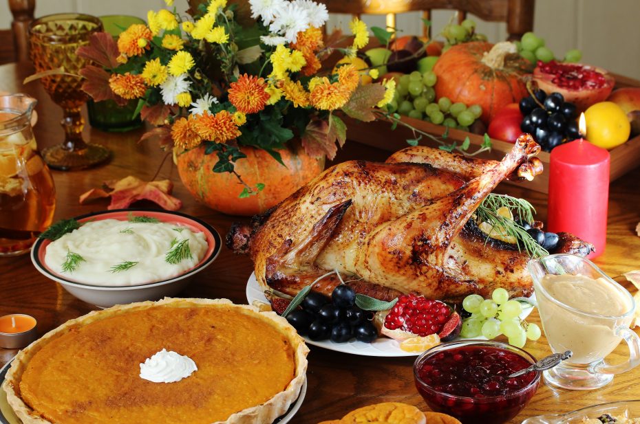 A Thanksgiving table featuring a roasted turkey, mashed potatoes, pumpkin pie, cranberry sauce, gravy, grapes, a floral centerpiece, and various other fruits and decorations.