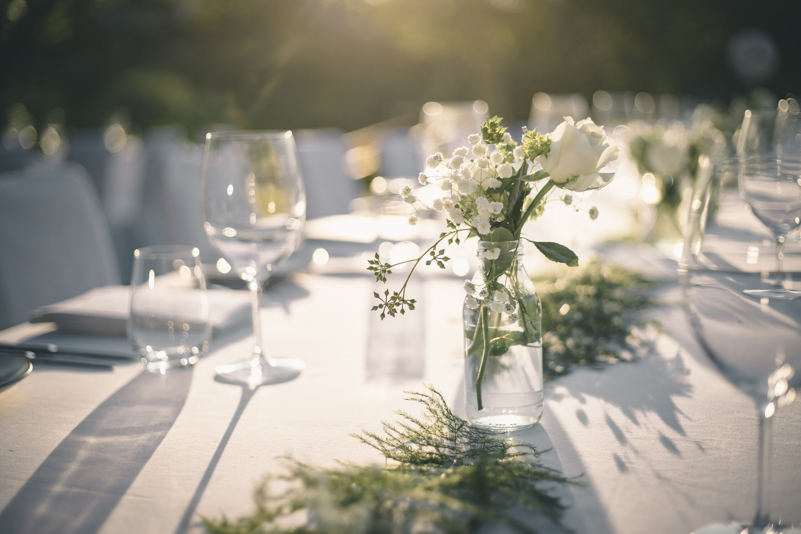 A table set for an outdoor event is adorned with clear glass vases containing white flowers, surrounded by empty wine glasses and greenery, bathed in sunlight.