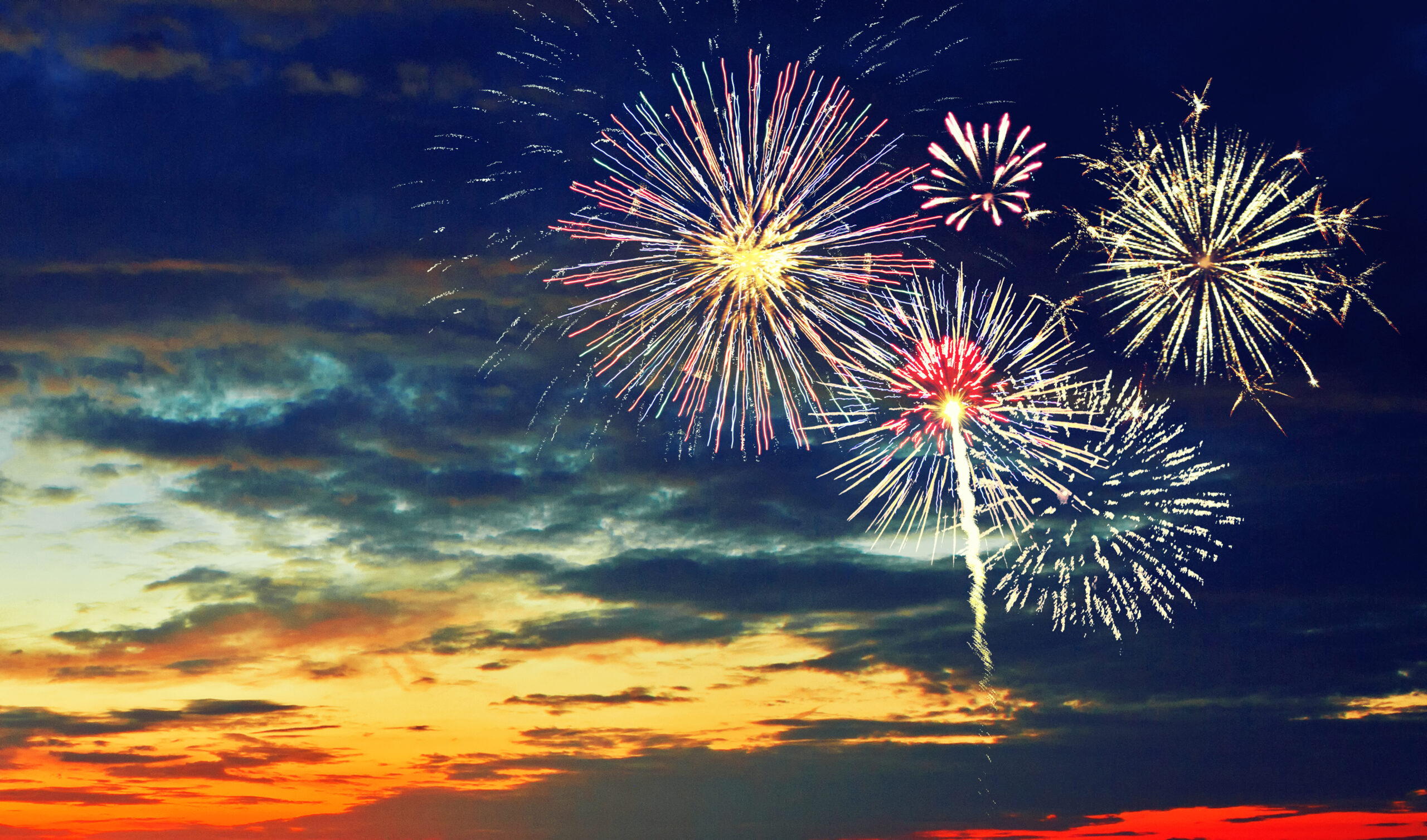 A dark sky illuminated by colorful fireworks bursts, with hues of red, blue, and yellow, against a backdrop of evening clouds and an orange-tinted horizon.