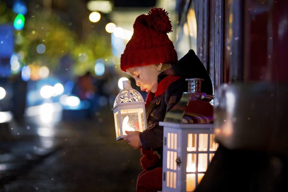 A child wearing a red hat and coat sits on a ledge holding a glowing lantern, with another lantern beside them. The background features blurred city lights at night.