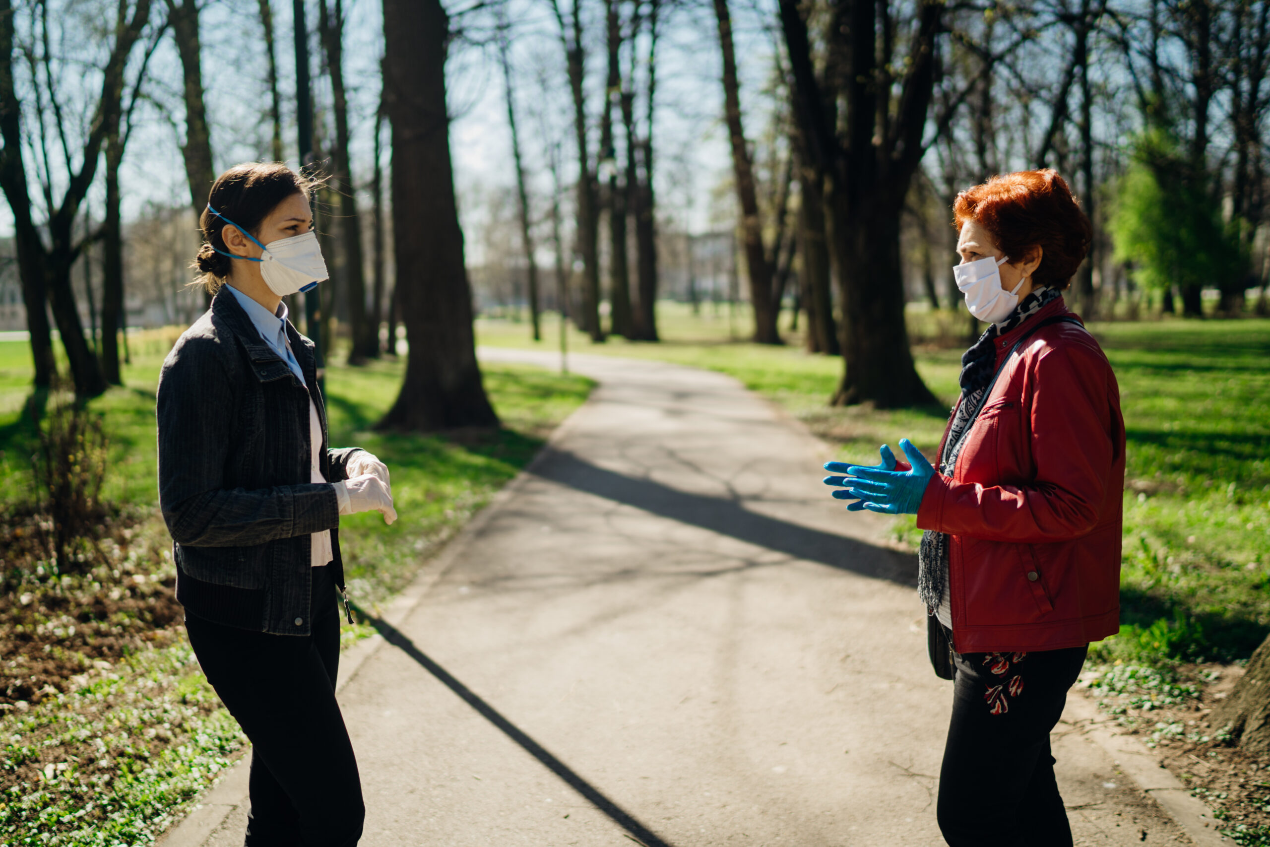 Two women stand on a park pathway wearing face masks and gloves, engaged in conversation. Trees and grass are visible in the background.