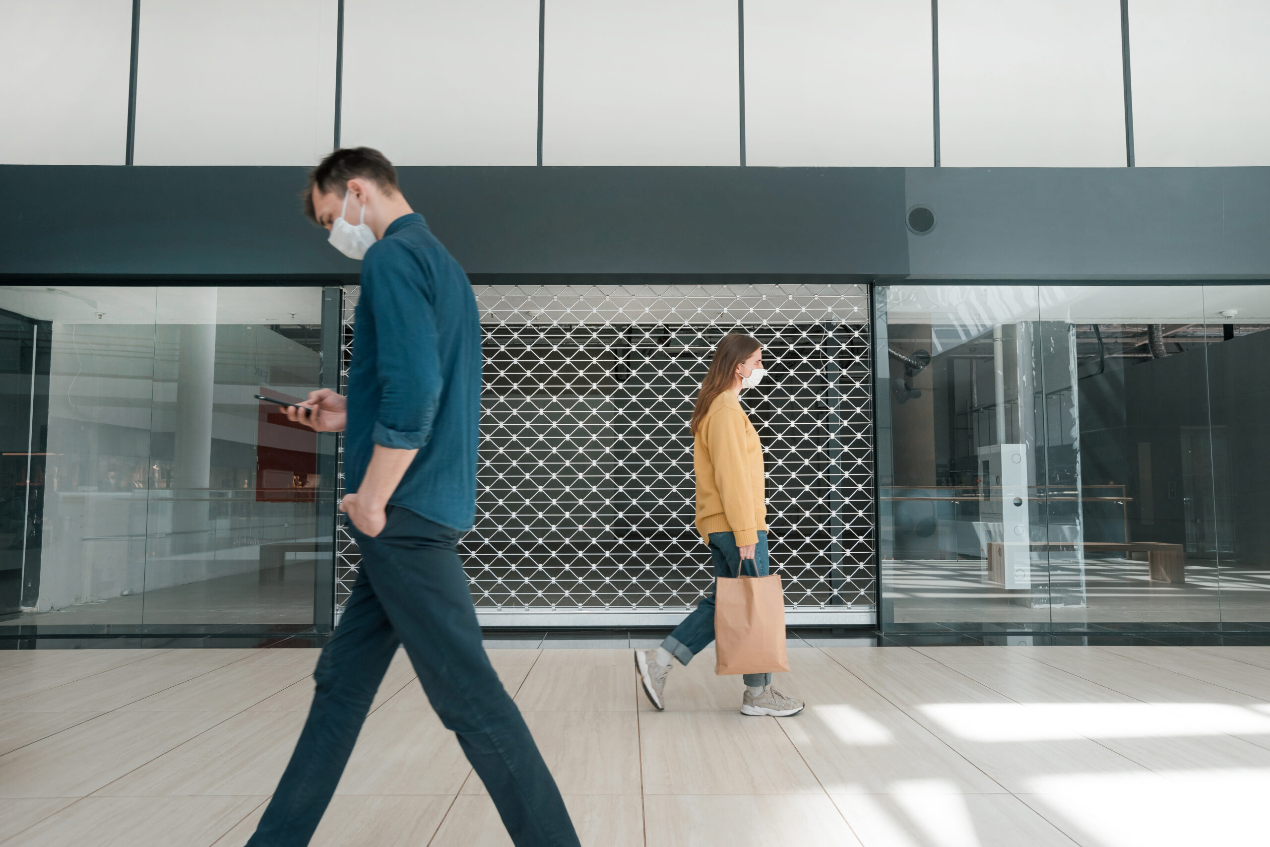 Two people wearing masks walk in a shopping mall, one carrying a paper bag. The shutters of a store are closed in the background.