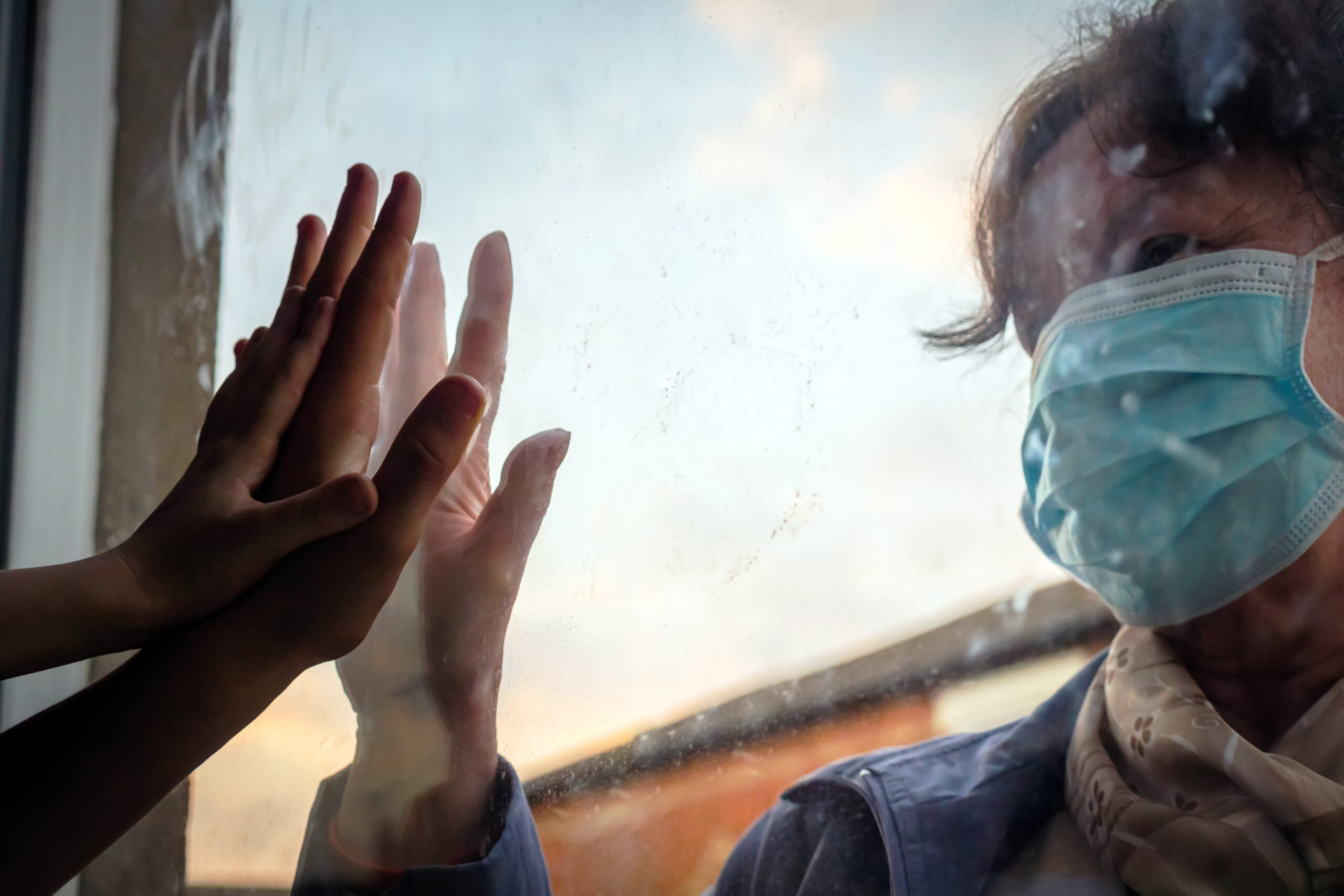 Two people touch their hands on opposite sides of a glass window, one wearing a blue surgical mask and a scarf.