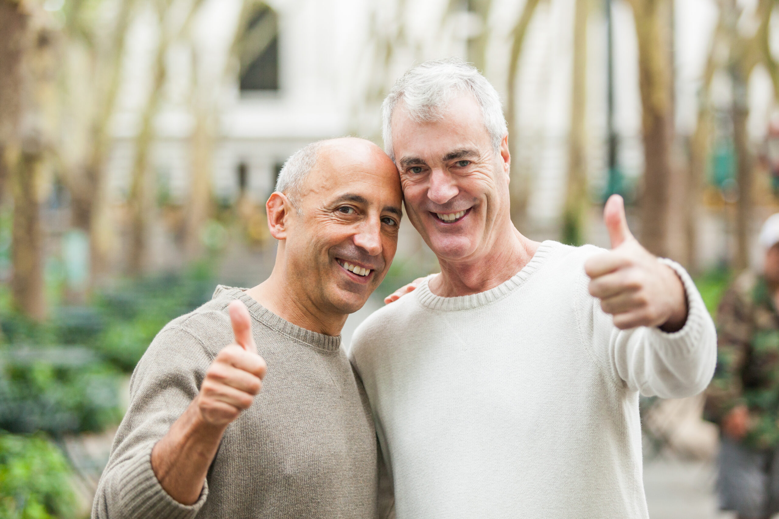 Two men, one with a grey sweater and the other with a tan sweater, smile and give thumbs up while standing outside. The setting, with trees and blurry background scenery visible behind them, captures a moment of camaraderie embodying Diversity, Equity, and Inclusion.