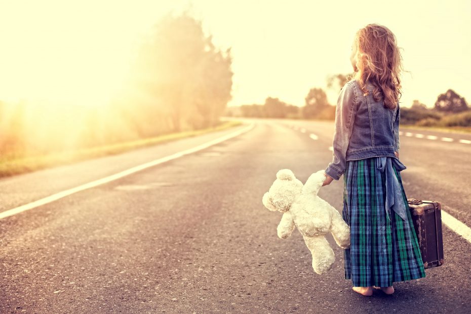 A child holding a teddy bear and a suitcase stands on an empty road, facing towards the horizon under a bright sky.