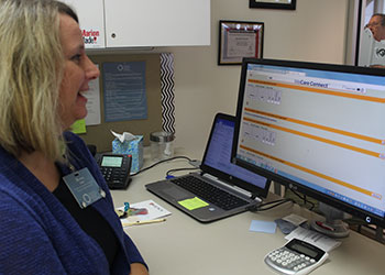 A woman is standing at a desk, looking at a computer screen. A laptop, calculator, and other office supplies are on the desk. Another person is partially visible in the background.