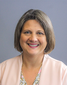 A woman with short brown hair, wearing a light pink blazer over a patterned blouse, smiles at the camera in front of a plain gray background.