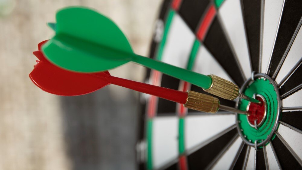 Close-up of a dartboard with two darts, one red and one green, embedded in the bullseye area. The dartboard's segmented sections are visible in the background.