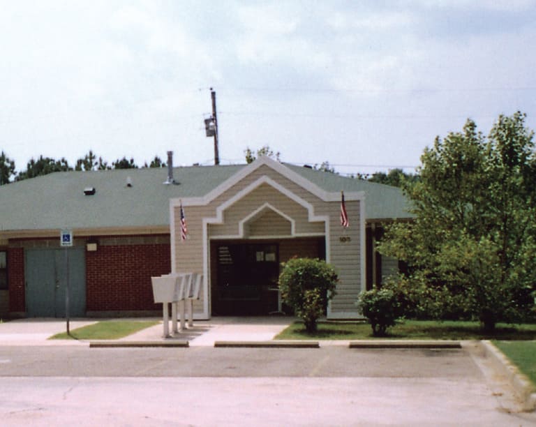 A single-story building with a green roof and brick facade stands proudly in Tupelo, Mississippi. Two American flags flank the front entrance, while mailboxes are situated nearby. Trees and a parking lot are visible in the foreground, embodying the charm of affordable housing in this area.