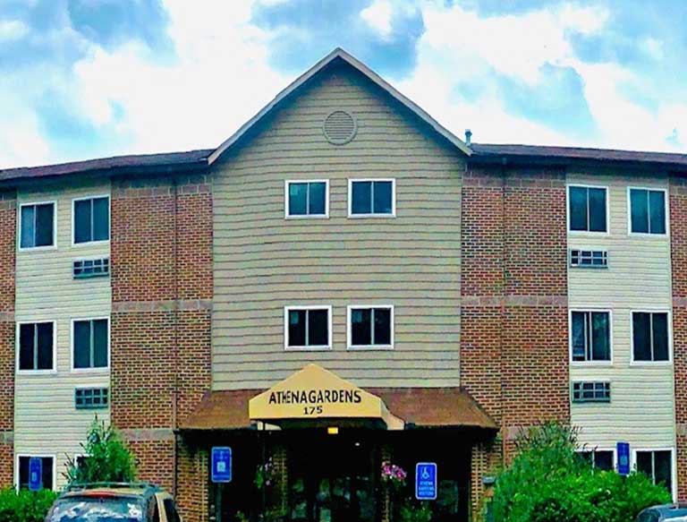 A three-story building with a sign that reads "Atena Gardens 175" above the entrance. The building, potentially one of the affordable housing options in Athens, Georgia, features a mix of brick and beige siding, several windows, and a nearby parking area.