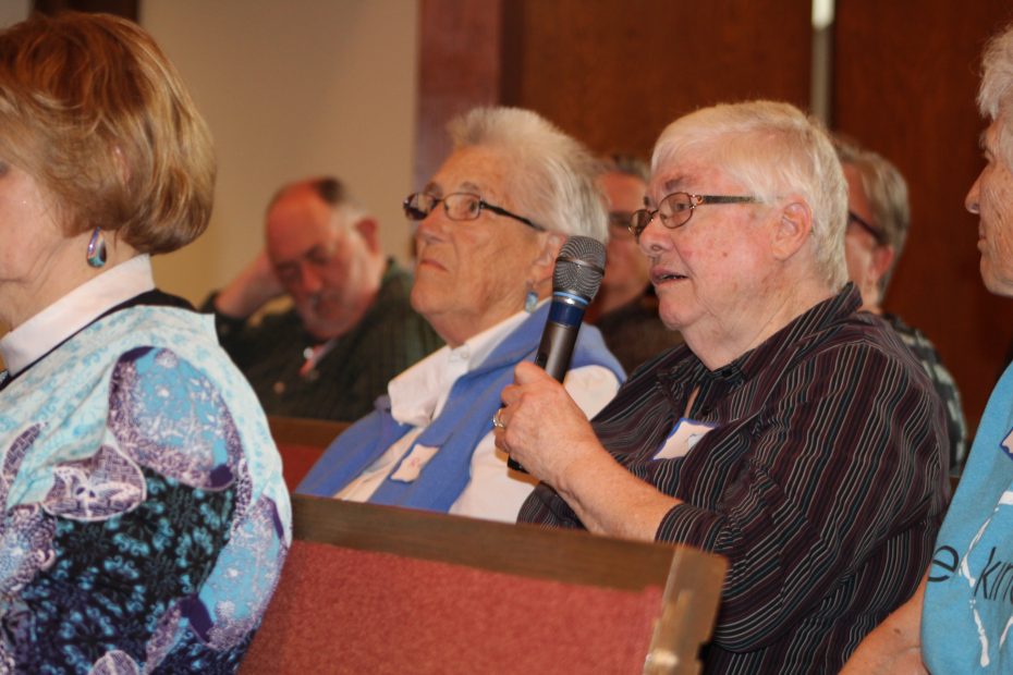 Several people are seated, and one individual is speaking into a microphone during an indoor event or meeting.