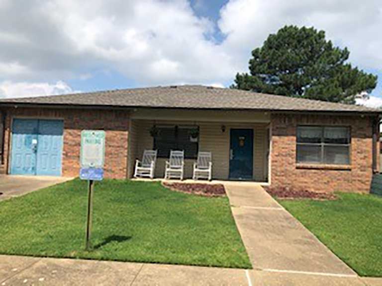 A one-story brick house with a gray roof, blue garage door, blue front door, and a small front porch with white rocking chairs. A sign in the front yard next to a walkway indicates affordable housing in Horn Lake, Mississippi.