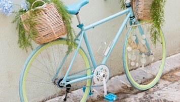 A pastel blue bicycle with yellow-green wheels, two wicker baskets on the front and back filled with green plants, is leaning against a light-colored wall.