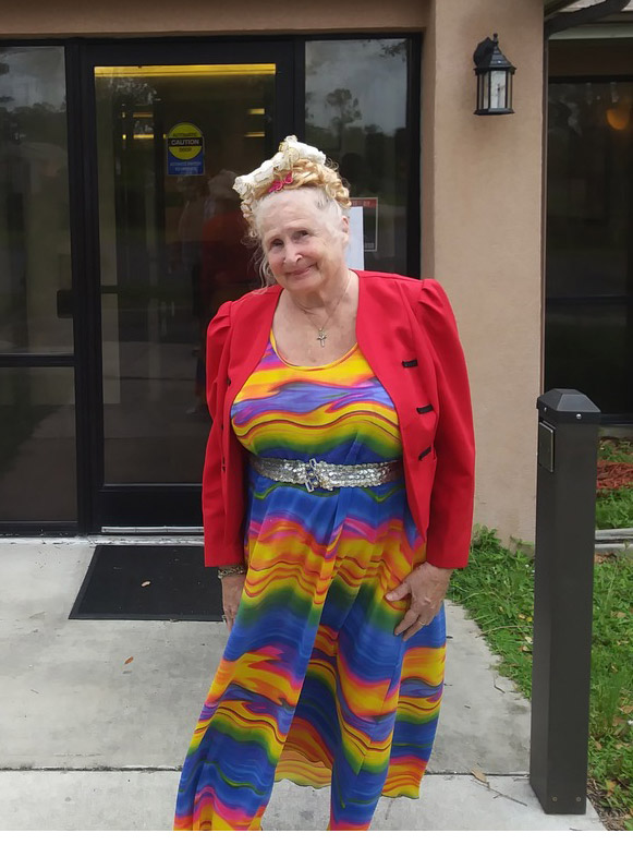 An elderly woman with gray hair stands outside a building, wearing a bright multicolored dress, a red jacket, and a jeweled belt. She has a flower hair accessory and smiles at the camera.