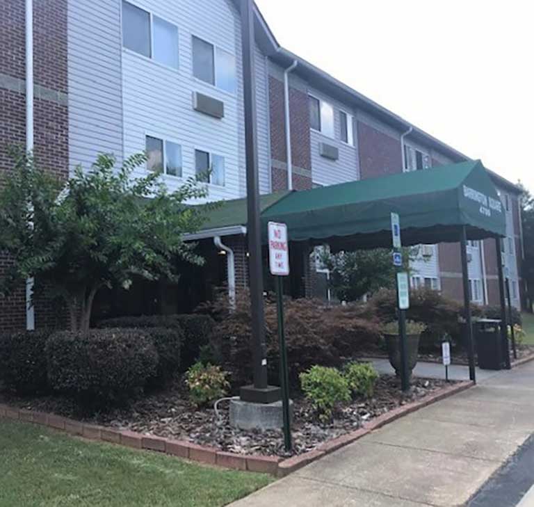 A multi-story brick and white-paneled building with a green canopy over the entrance stands proudly as part of the affordable housing in Acworth, Georgia. Shrubs and a sidewalk are in the foreground, while a signpost nearby reads "No Parking Fire Lane.