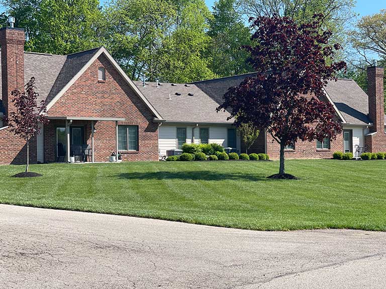 A brick house with multiple peaked roofs, surrounded by neatly trimmed grass and young trees. A paved road runs in front of the house, with leafy trees and shrubs visible in the background.