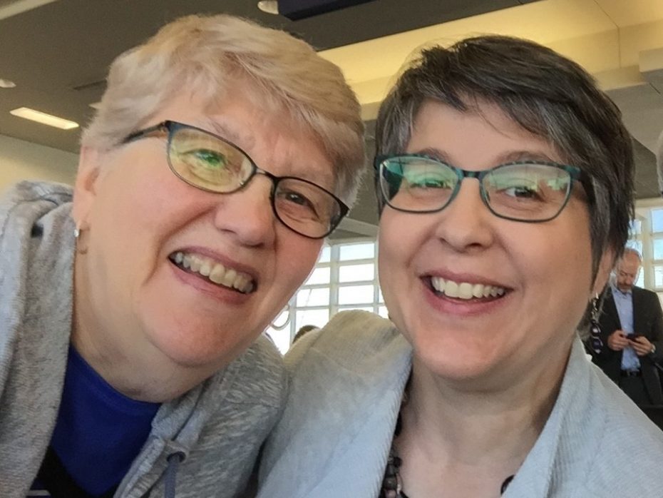 Two smiling women, both wearing glasses, are taking a selfie indoors. One has short blond hair, and the other has short gray hair. The background shows a modern interior setting with another person in the distance.