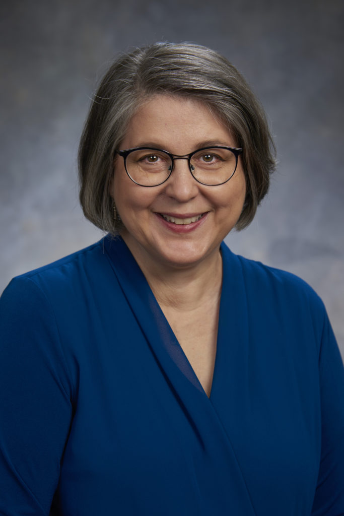 A woman with short gray hair and glasses, embodying leadership, wears a blue blouse and smiles while posing for a portrait against a neutral background.