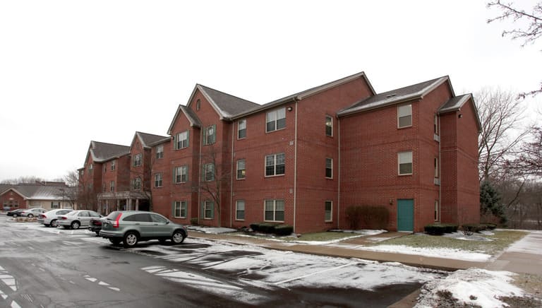 Three-story red brick apartment building with several windows on each floor, offering affordable housing in Columbus, Ohio. Snow patches are visible on the ground and parked cars line the street in front. Trees without leaves are seen in the background.