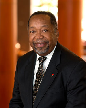 Man in a black suit and patterned tie sits and smiles at the camera. Brown wooden structures are blurred in the background.