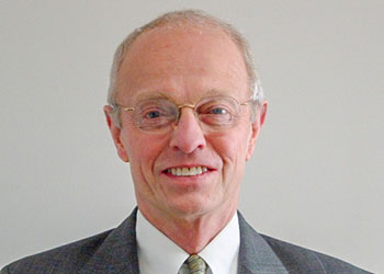 A man wearing glasses and a gray suit with a white shirt and tie, posing against a plain background.