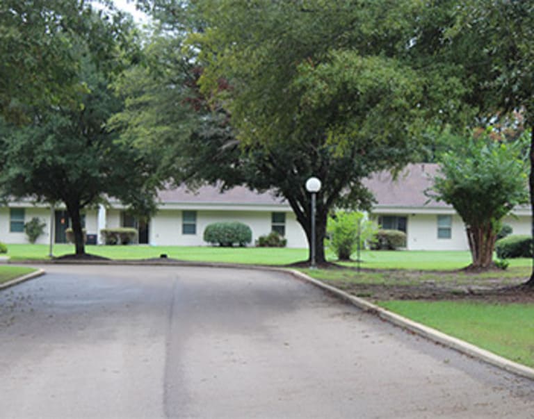 A paved road curves toward single-story white buildings surrounded by green lawns and trees, offering a picturesque view of affordable housing in Starkville, Mississippi.