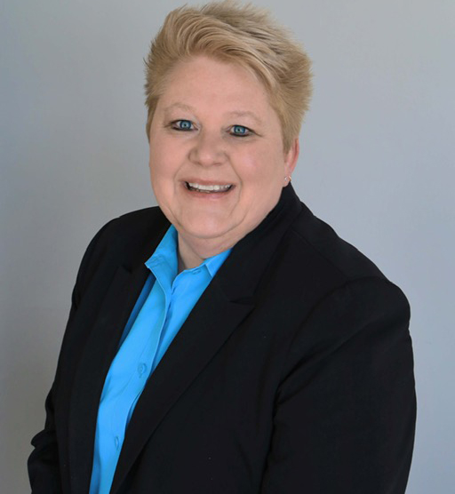 A person with short blond hair wearing a black blazer and blue shirt smiles while posing against a plain background.