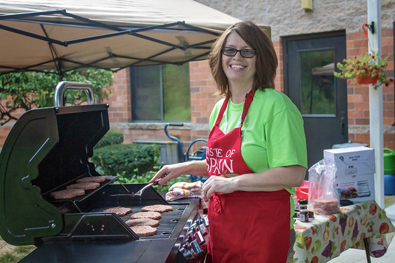 A woman smiles while grilling burgers under a canopy, wearing a bright green shirt and a red apron that says "Taste of Japan." With determination in her eyes, she is on a mission to deliver the best flavors. A table with various items is in the background.