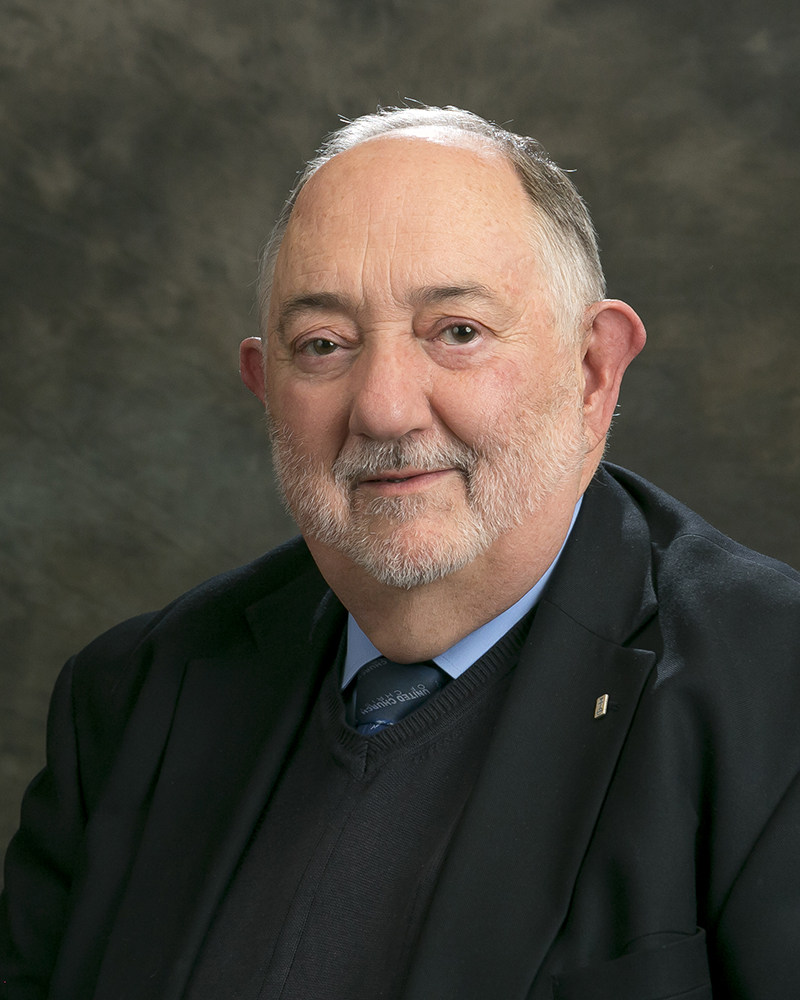 An older man with a gray beard and short hair is wearing a suit and tie, posing for a professional portrait against a neutral background.