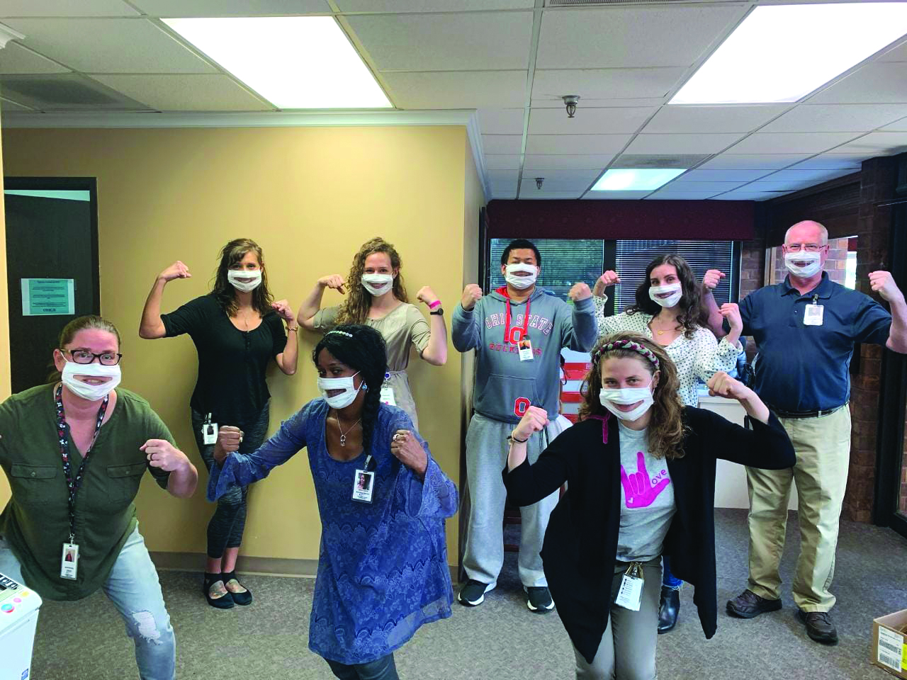 A group of eight people standing in an office space, all wearing partially transparent face masks, posing with flexed arms. The team proudly supports Columbus Colony Elderly Care, showcasing their commitment and strength.