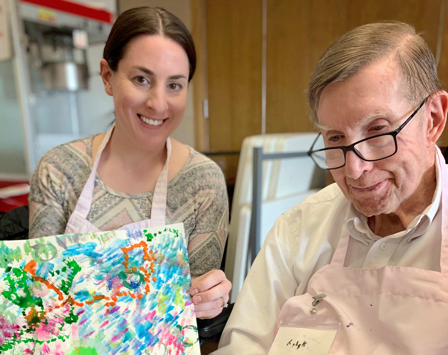 A woman and an elderly man sit together; the woman holds up a colorful piece of artwork, both are wearing aprons.